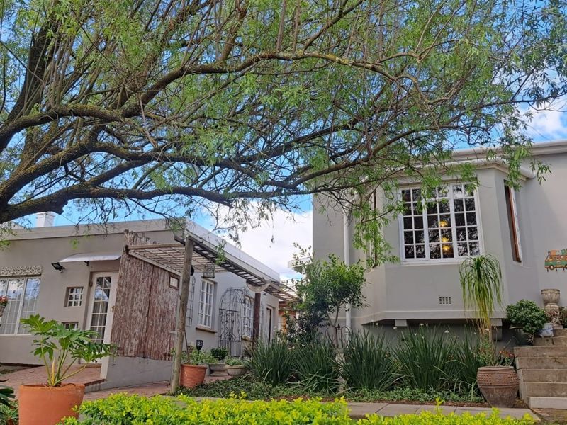 A charming rear exterior view featuring a bay window, paved patio area, pergola, and landscaped garden with concrete steps leading to the entrance.