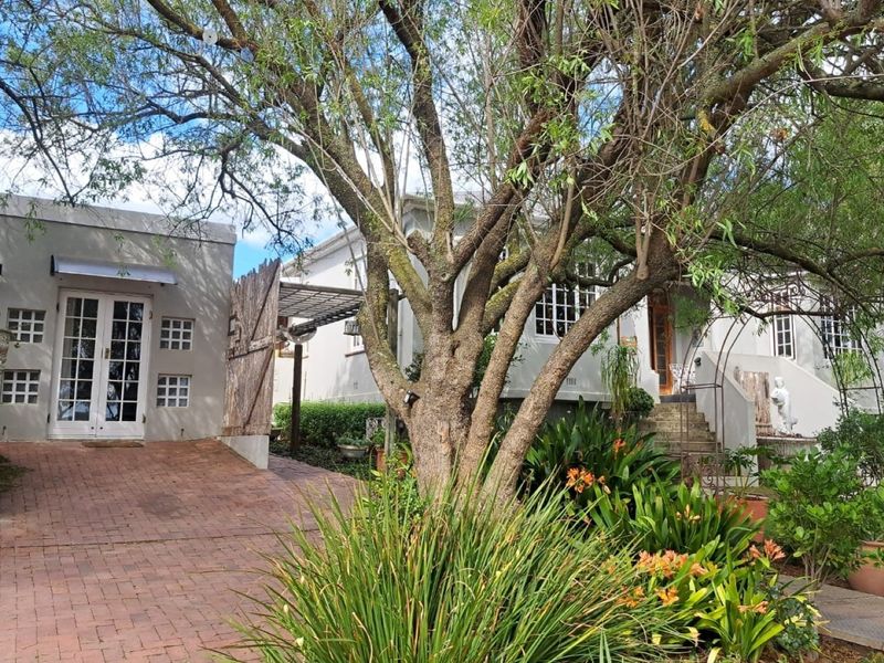 Front exterior view of the property featuring a brick paved driveway, mature trees, landscaped garden, and access to the main entrance and side structure.