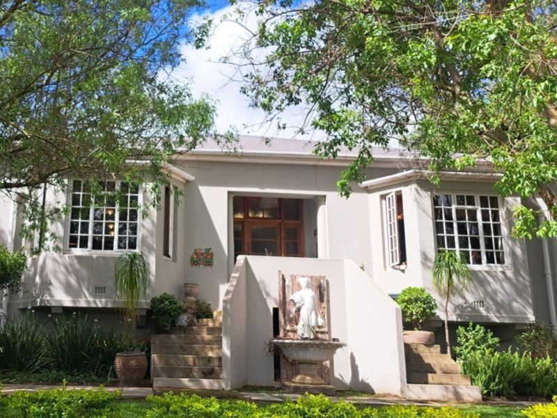 A charming front facade of a house featuring a central staircase leading to the entrance, flanked by multi-pane windows and mature garden landscaping.