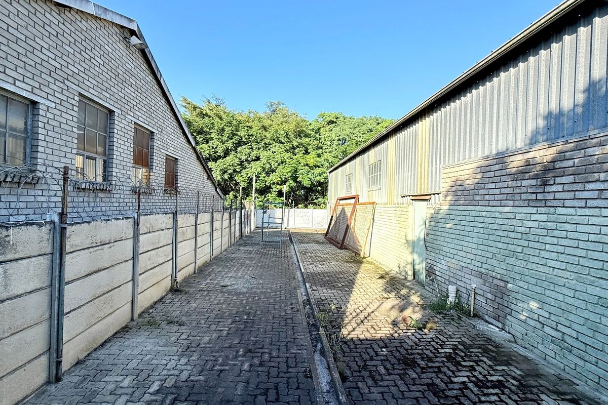 A narrow exterior side passage between two industrial buildings featuring paved ground, brick perimeter walls with security fencing, and metal cladding.