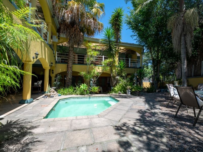 A sunny outdoor pool area featuring stone paving, a small swimming pool, and a two-story lodge structure with covered patios and balconies.