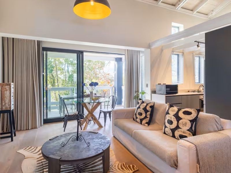 Bright open-plan living area featuring hardwood floors, exposed ceiling beams, and seamless flow to a private balcony, complemented by an integrated kitchen space.