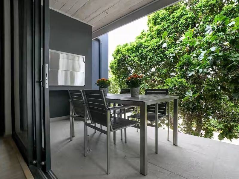 A covered balcony featuring concrete flooring, folding glass doors, and a built-in stainless steel braai, overlooking lush greenery.