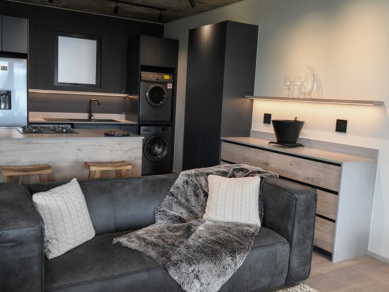 Modern open-plan living area featuring a sleek kitchen with integrated appliances, dark cabinetry, a central island, and contemporary concrete ceiling finishes.