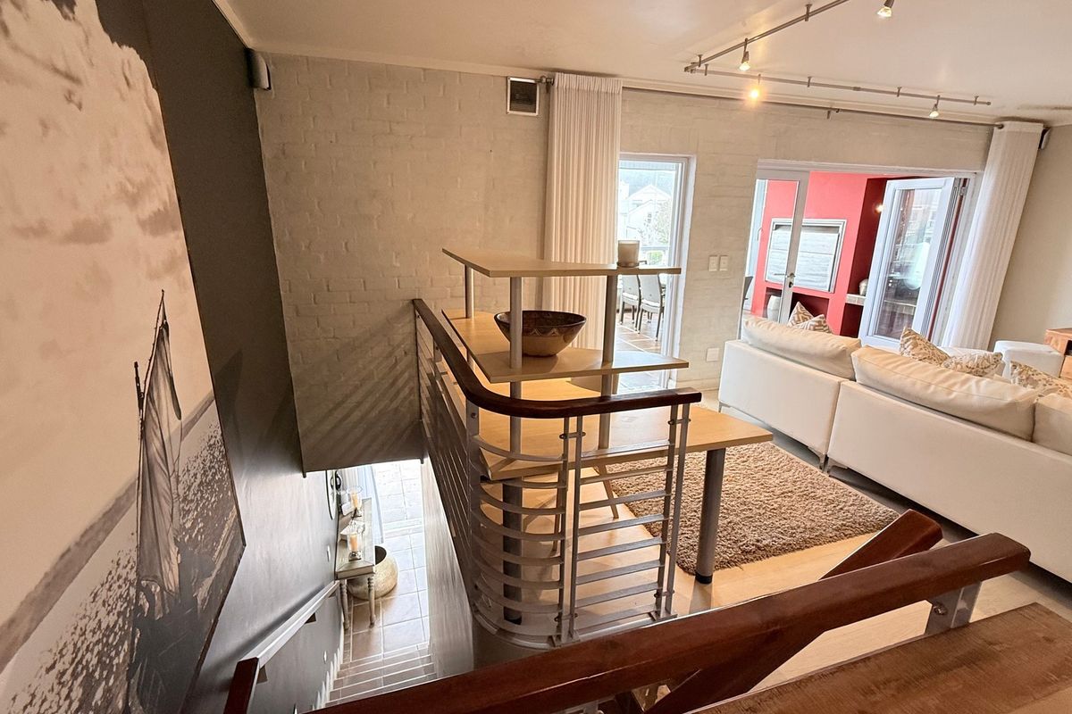 A bright living area featuring a staircase with a metal balustrade, exposed brick walls, and direct access to an outdoor patio through glass sliding doors.