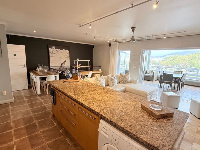 Spacious open-plan living area featuring a large granite kitchen island, integrated laundry appliance, track lighting, and seamless flow to an outdoor balcony.