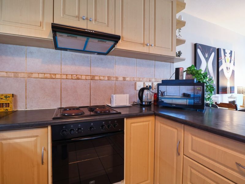 A functional kitchen featuring light wood cabinetry, dark laminate countertops, a tiled backsplash, and integrated cooking appliances including an oven and extractor fan.