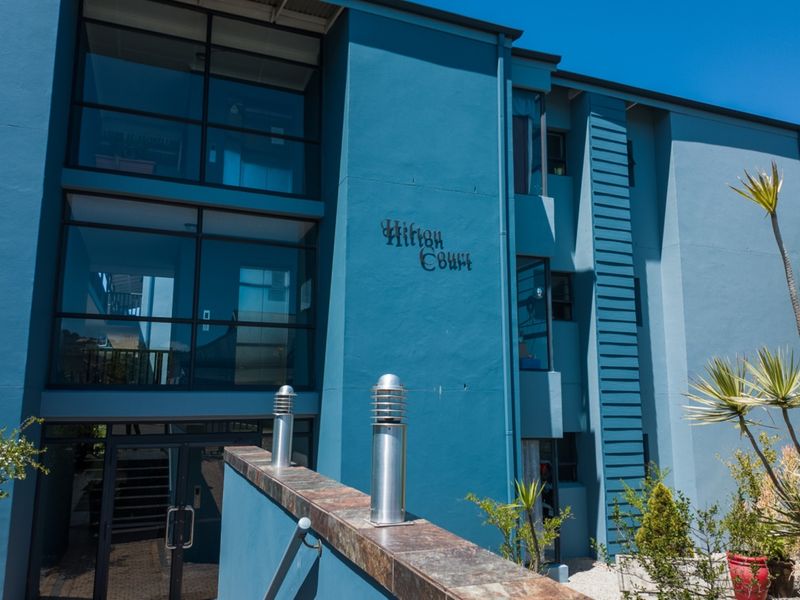 Front facade of the Hilton Court apartment building featuring modern blue exterior paint, large glass windows, and landscaped entrance area.