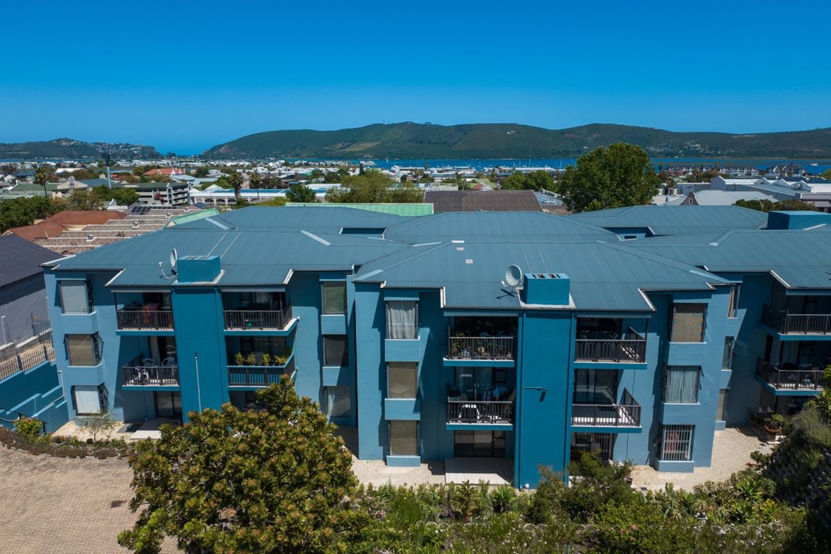 Aerial view of a multi-story apartment complex featuring blue exterior walls, metal roofing, and private balconies with scenic coastal and mountain views.