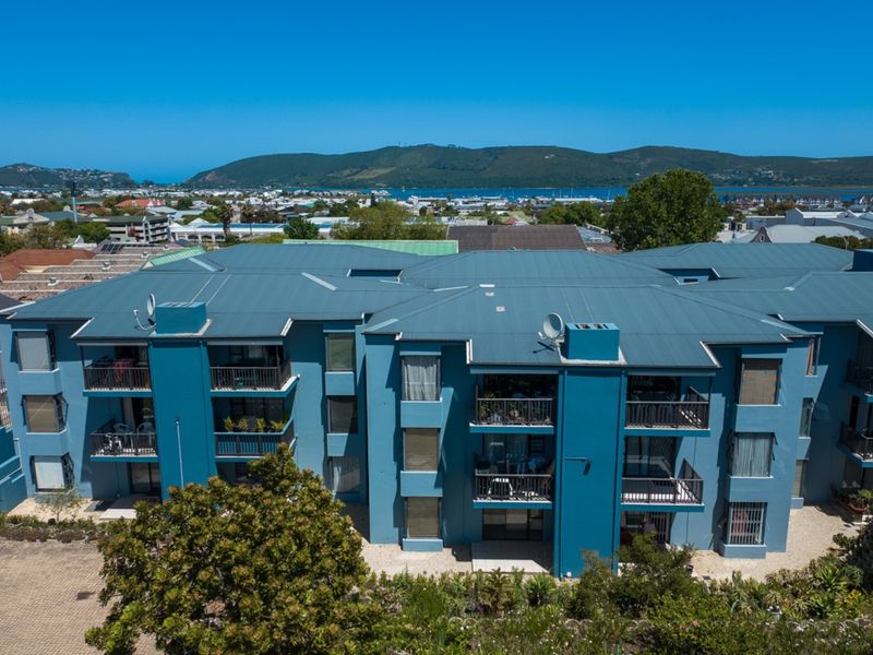 Aerial view of a multi-story apartment complex featuring blue exterior walls, metal roofing, and private balconies with scenic coastal and mountain views.