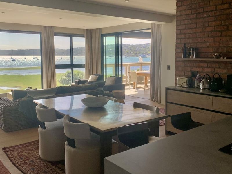 Spacious open-plan living and dining area featuring an exposed brick wall, modern cabinetry, and large sliding doors opening to a scenic waterfront balcony.