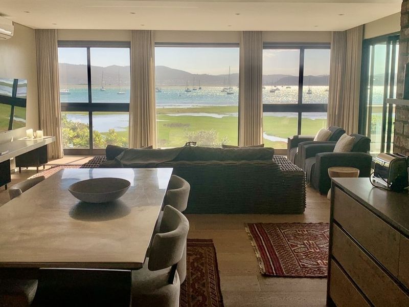 Spacious open-plan living and dining area featuring exposed brick accents, stone flooring, and expansive floor-to-ceiling windows offering stunning panoramic water and landscape views.