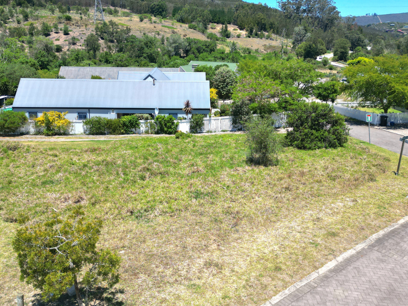 An elevated view of a vacant residential plot featuring established grass, a paved driveway, and surrounding white picket fencing in a scenic neighborhood.