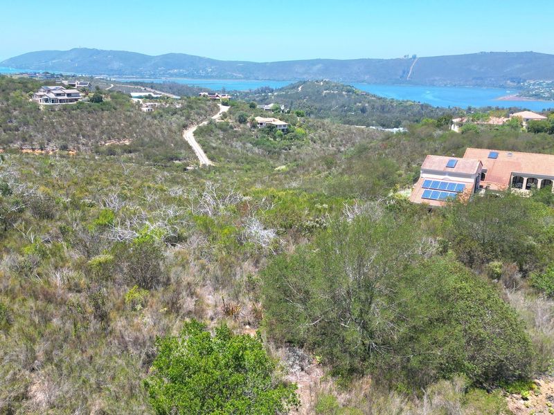 An aerial view of a vacant residential plot featuring natural vegetation and sloped terrain with surrounding landscape and water views.