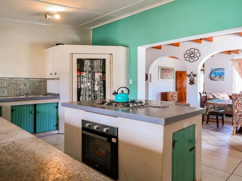 Open-plan kitchen featuring a central island with built-in oven and hob, flowing into an adjacent dining area with exposed timber ceiling beams.