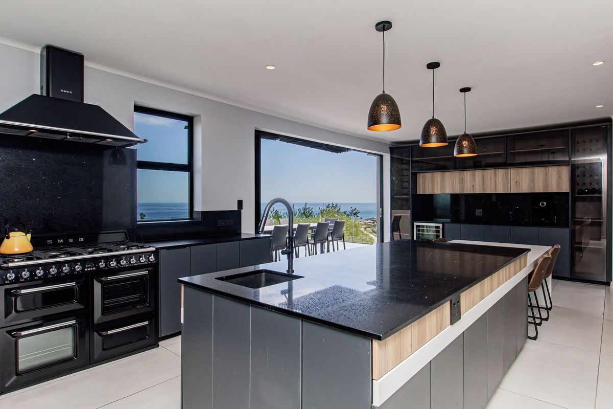 Modern open-plan kitchen featuring a large island, premium black cabinetry, stone countertops, high-end gas range, and seamless access to an outdoor patio with ocean views.