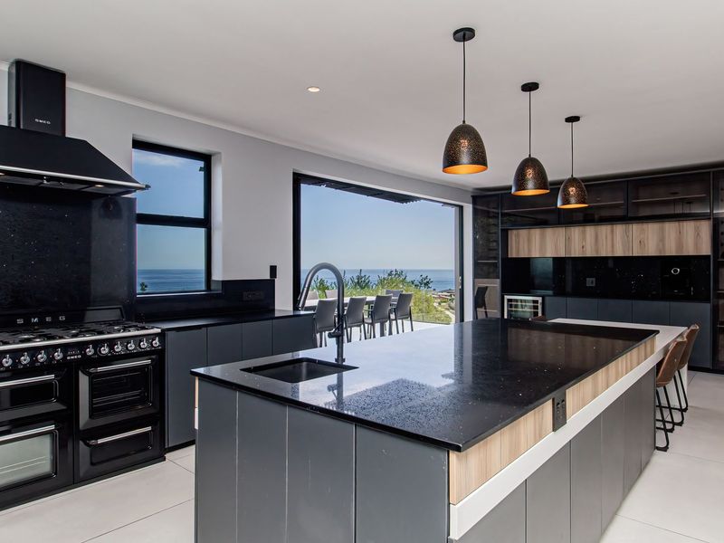 Modern open-plan kitchen featuring a large island, premium black cabinetry, stone countertops, high-end gas range, and seamless access to an outdoor patio with ocean views.