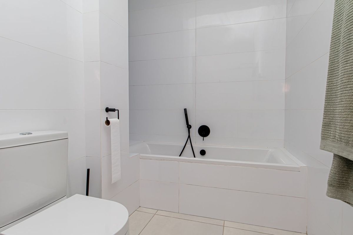 A modern, minimalist bathroom featuring white wall and floor tiles, a built-in bathtub with matte black fixtures, and a clean, contemporary design.