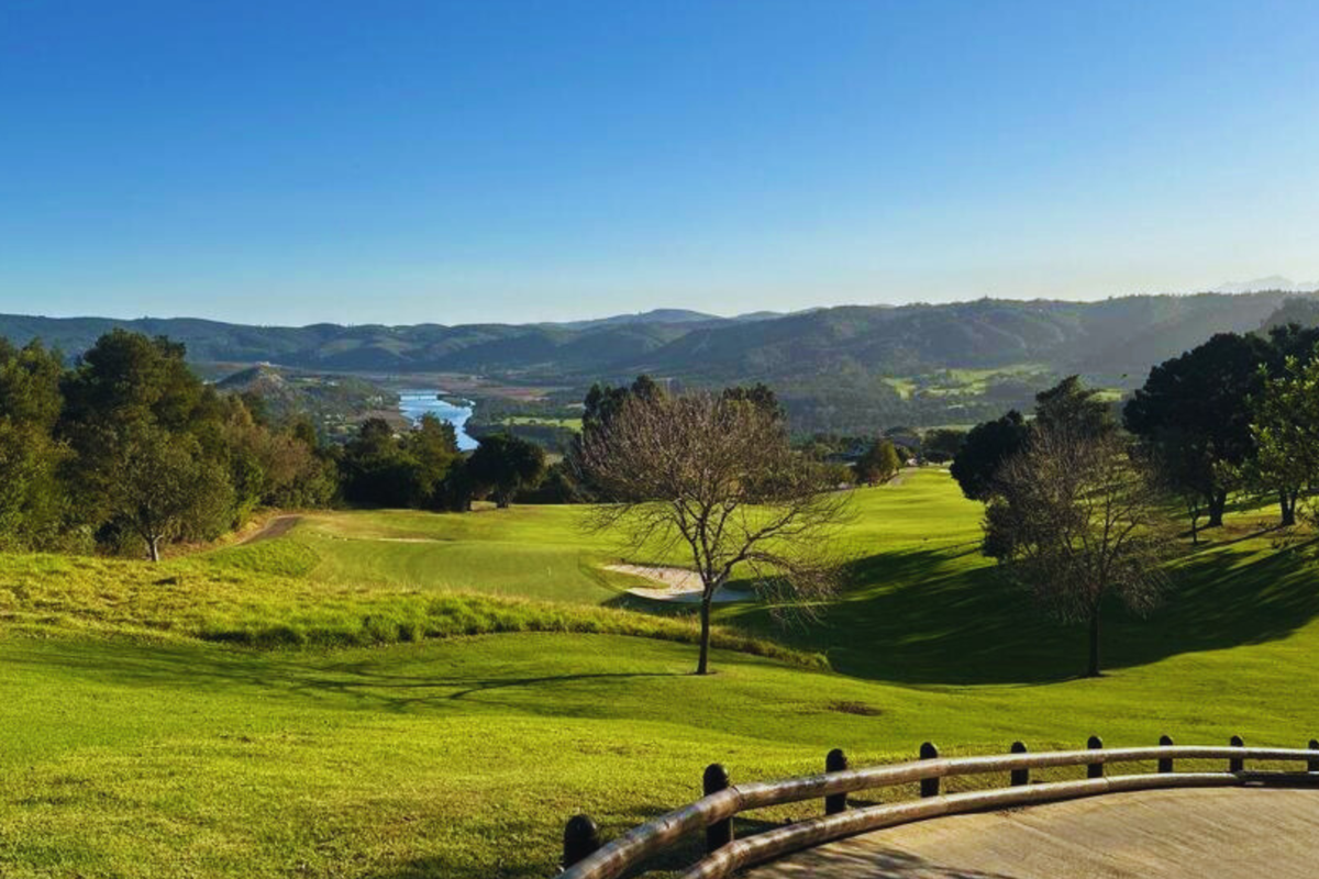 A scenic elevated view overlooking a lush green golf course, rolling hills, and a distant river, framed by a timber rail fence.