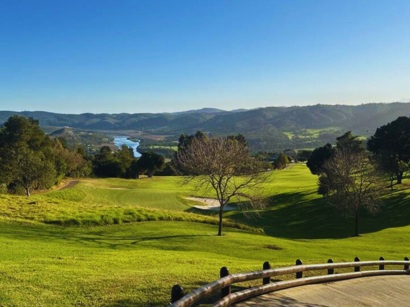A scenic elevated view overlooking a lush green golf course, rolling hills, and a distant river, framed by a timber rail fence.