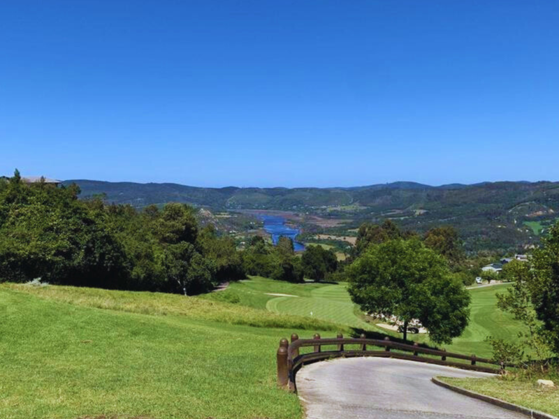 A scenic elevated view overlooking a lush green landscape, golf course fairways, and a distant river valley under a clear blue sky.