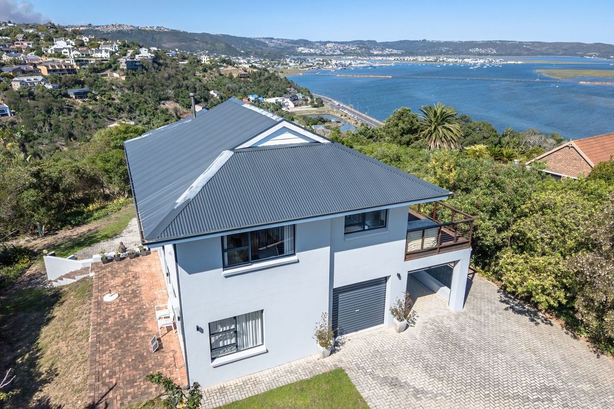 Aerial view of a two-story house featuring a corrugated metal roof, paved driveway, garage, and balcony, set against a scenic coastal landscape.
