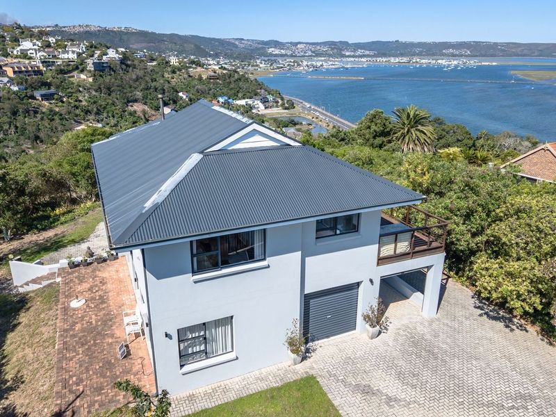 Aerial view of a two-story house featuring a corrugated metal roof, paved driveway, garage, and balcony, set against a scenic coastal landscape.