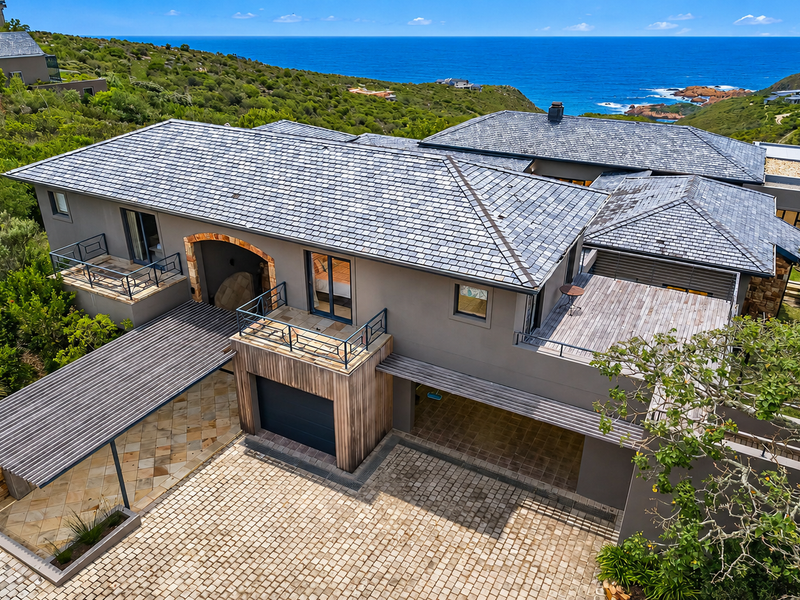 Aerial view of a modern coastal residence featuring a slate roof, stone accents, timber decking, and a paved driveway overlooking the ocean.