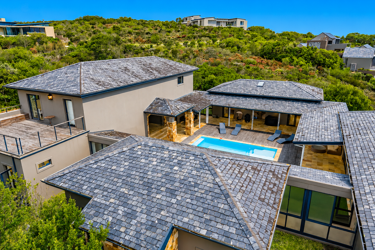 Aerial view of a modern residence featuring a central swimming pool, expansive timber decking, and distinctive slate roof tiles set within a natural landscape.