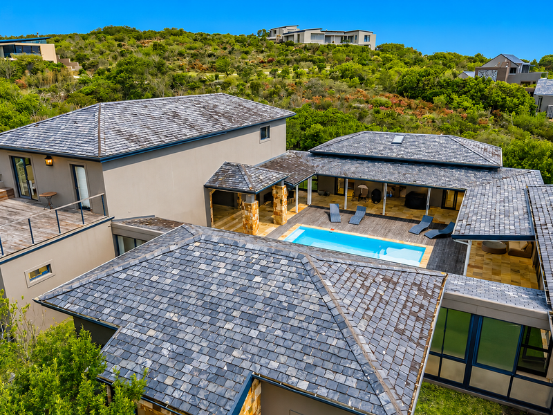 Aerial view of a modern residence featuring a central swimming pool, expansive timber decking, and distinctive slate roof tiles set within a natural landscape.