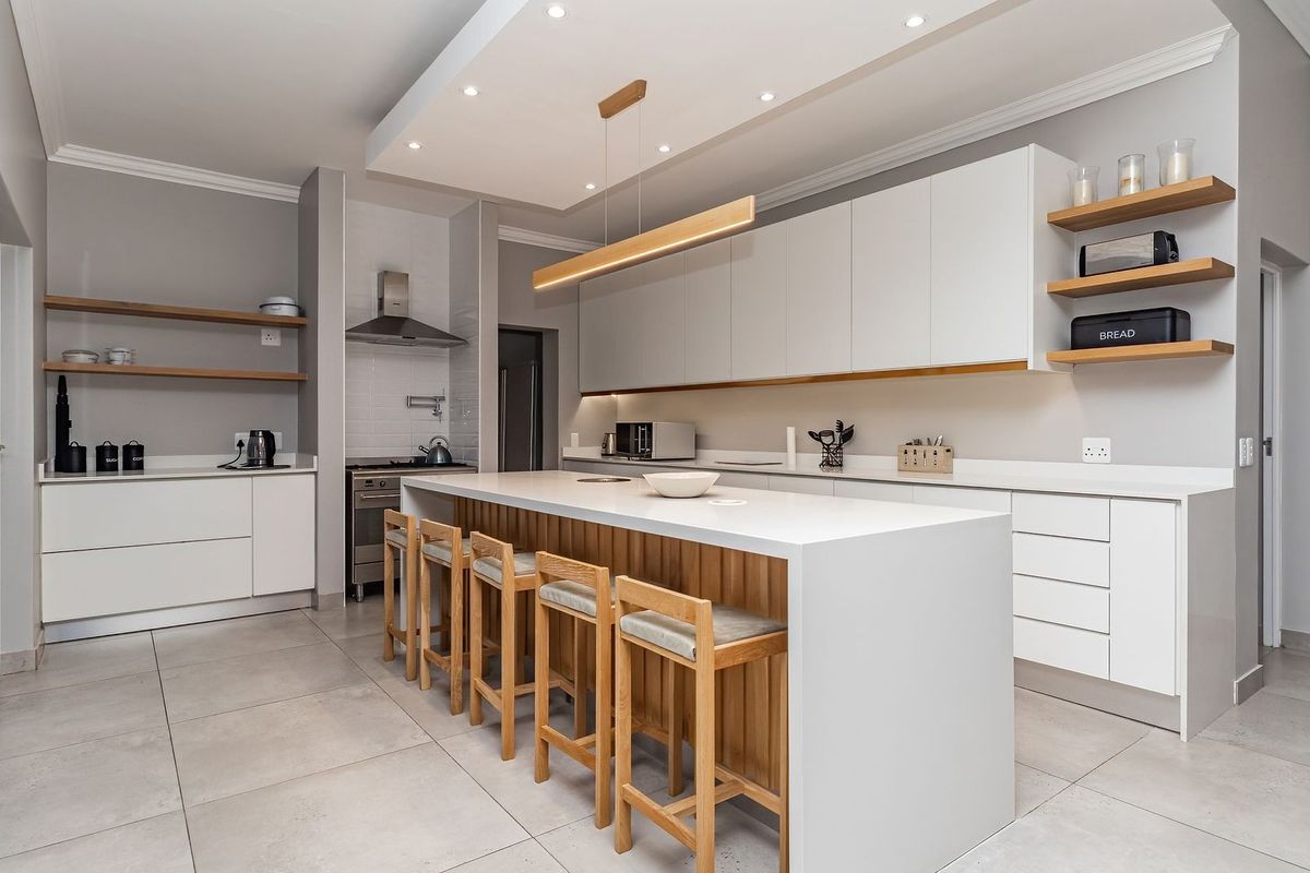 Modern, well-appointed kitchen featuring a large central island, sleek white cabinetry, stone countertops, and high-quality integrated lighting, opening onto an outdoor area.