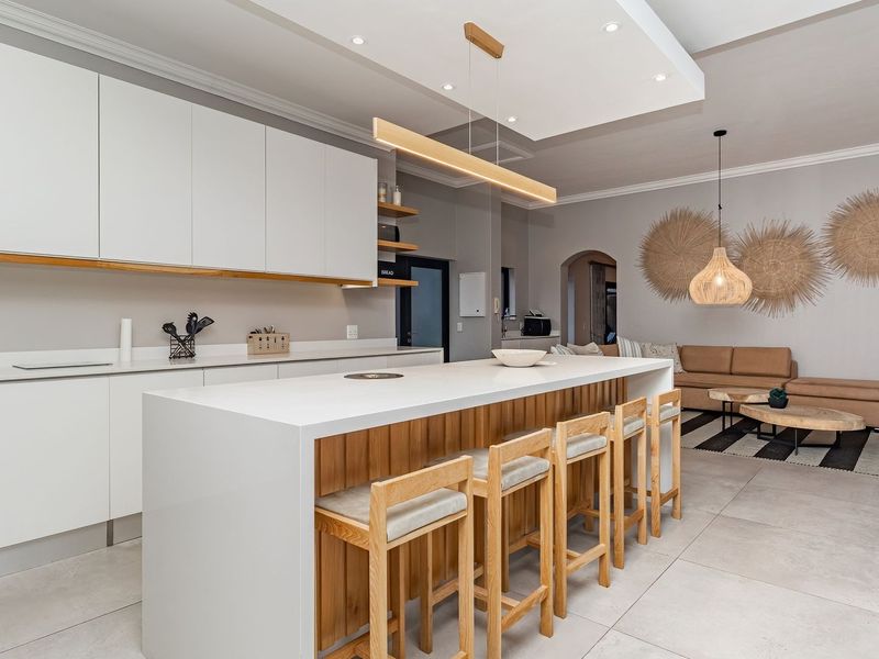 Modern open-plan kitchen and living area featuring a large central island, sleek white cabinetry, stone countertops, and contemporary recessed and pendant lighting.