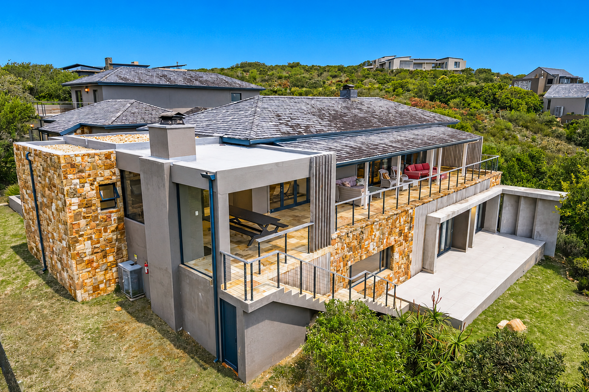 Aerial view of the rear elevation featuring stone cladding, a spacious tiled patio, a balcony with glass balustrades, and an external staircase.