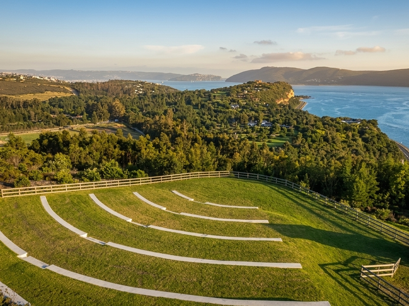 Elevated view of a landscaped outdoor amphitheater area featuring tiered concrete seating, timber fencing, and expansive coastal and forest scenery.