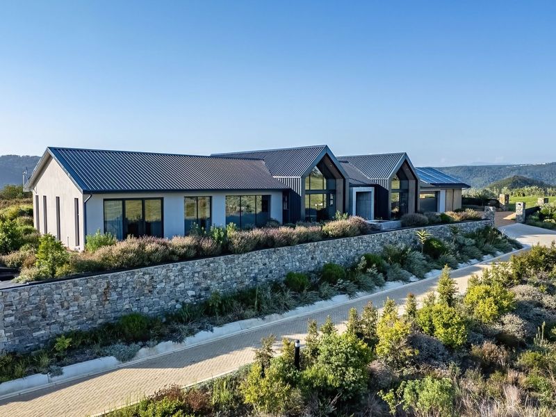 Aerial view of a modern residential property featuring a metal roof, stone retaining wall, and landscaped surroundings with a paved driveway.