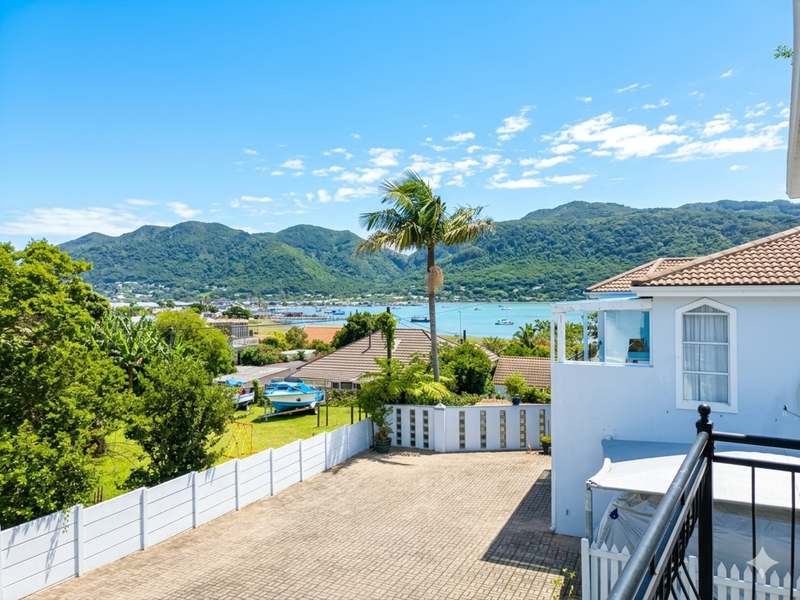 Elevated view overlooking a residential property with a paved driveway, white fencing, and scenic mountain and coastal water vistas in the background.