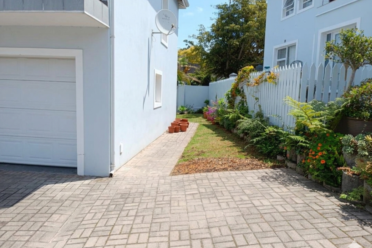 A side exterior view showing a paved driveway, garage access, and a landscaped garden area with a white picket fence.