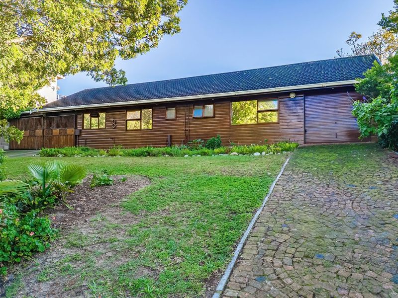 Front elevation of a single-story house featuring timber cladding, a tiled roof, and a cobblestone driveway set within a landscaped garden.