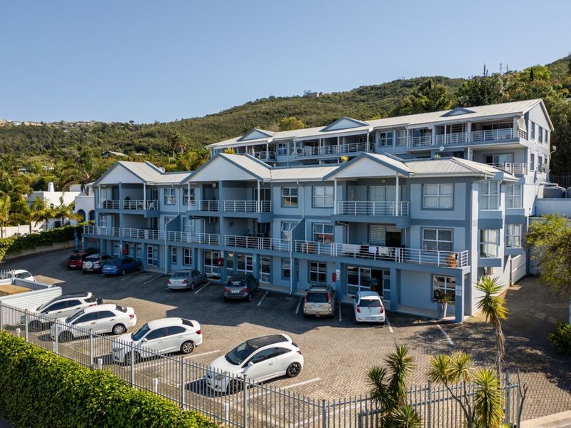 Aerial view of a multi-story apartment complex featuring paved parking, private balconies with metal railings, and a hillside backdrop.