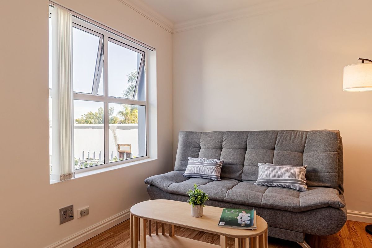 A bright living area featuring laminate flooring, neutral walls, and a large window with vertical blinds, providing natural light and a pleasant outlook.