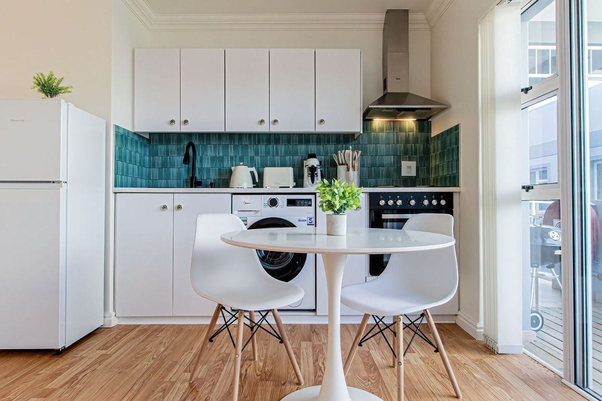 Compact modern kitchen featuring white cabinetry, a vibrant teal tiled backsplash, integrated washing machine, built-in oven, and direct access to an outdoor balcony area.