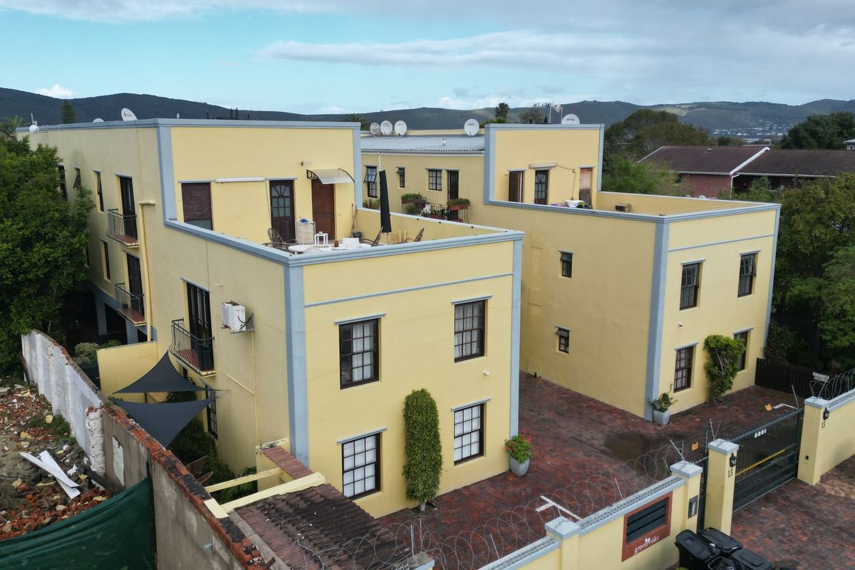 Aerial view of a multi-story apartment complex featuring yellow facades, paved driveway, security perimeter fencing, and rooftop terrace areas.
