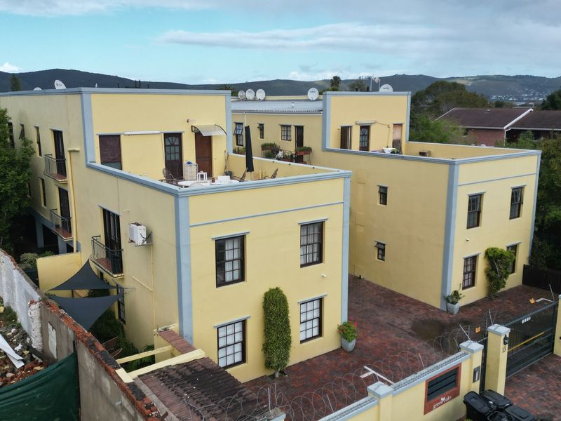Aerial view of a multi-story apartment complex featuring yellow facades, paved driveway, security perimeter fencing, and rooftop terrace areas.