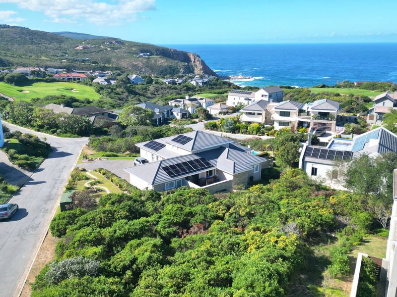 Aerial view of a residential coastal estate featuring homes with solar panels, surrounded by lush greenery and proximity to the ocean.