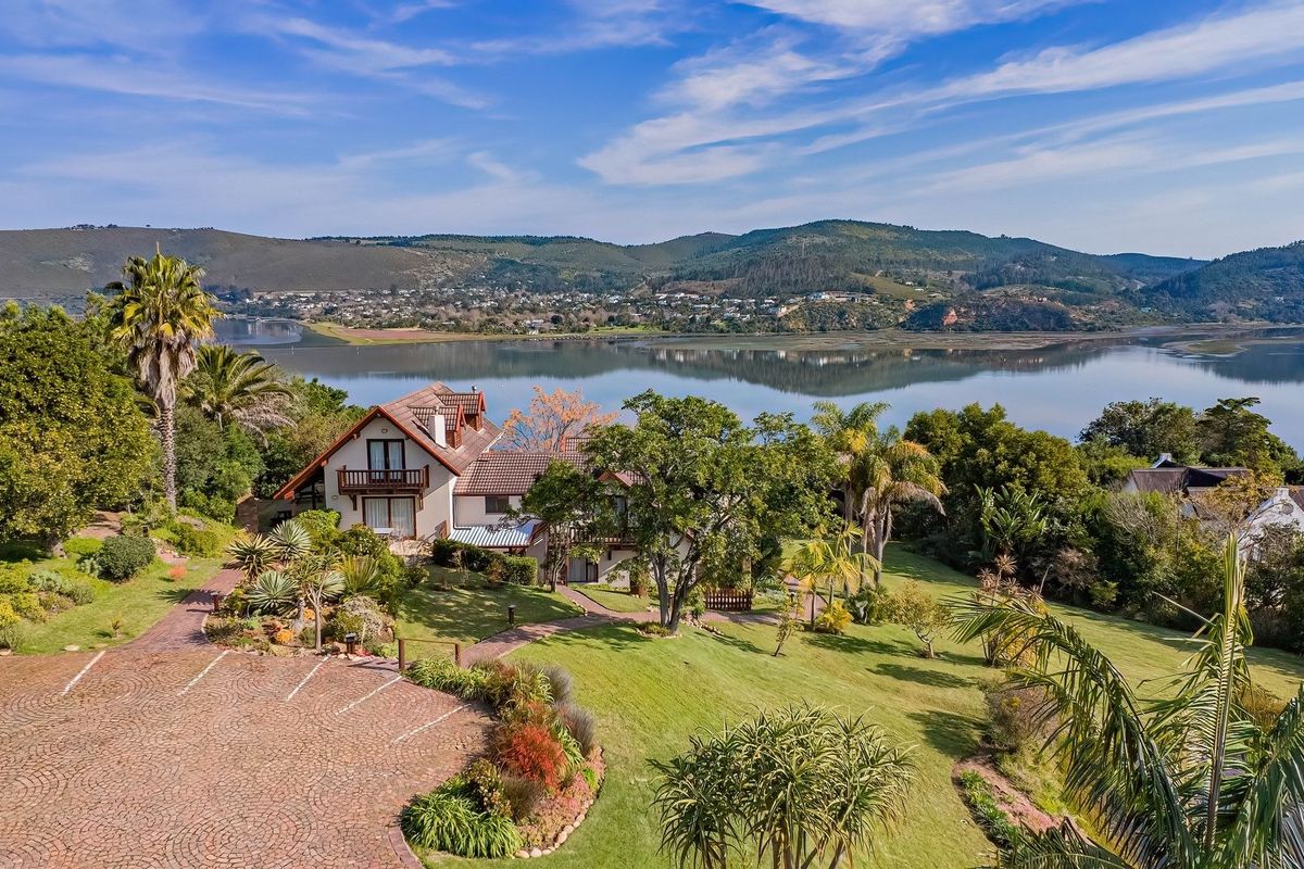 Aerial view of a lodge property featuring a paved driveway, expansive landscaped gardens, and a balcony overlooking a scenic lagoon and mountain landscape.