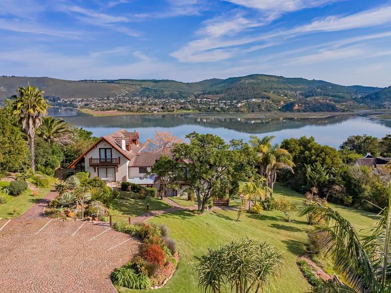 Aerial view of a lodge property featuring a paved driveway, expansive landscaped gardens, and a balcony overlooking a scenic lagoon and mountain landscape.