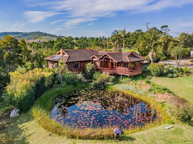 Aerial view of a timber lodge featuring a wrap-around deck, tiled roof with solar panels, and a scenic pond set within a landscaped garden.