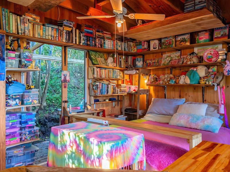 A cozy bedroom featuring warm timber wall cladding, exposed ceiling beams, a ceiling fan, and extensive built-in shelving with direct access to the outdoors.