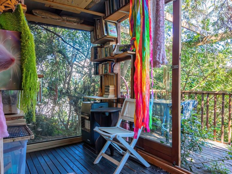 An elevated timber balcony or sunroom featuring large glass panels, timber decking, and a wooden balustrade overlooking surrounding trees.