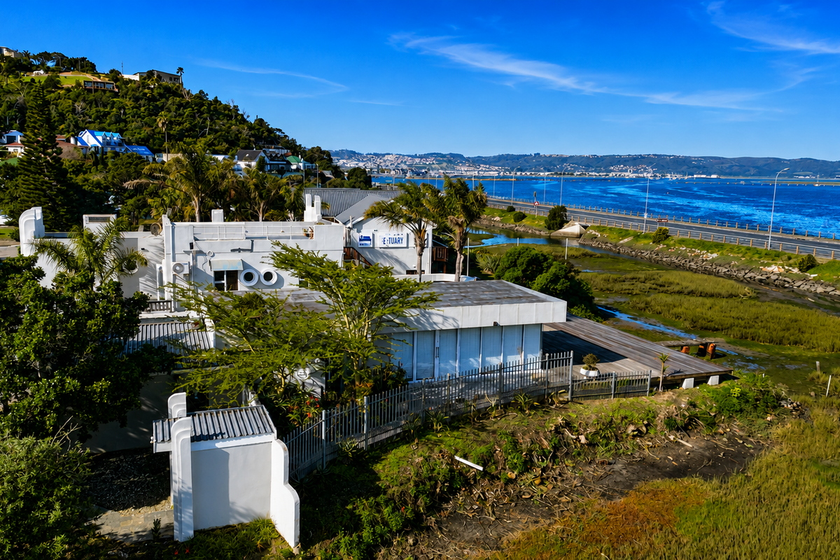 Aerial view of the property featuring a timber deck, metal fencing, and flat roof architecture, situated near the estuary and coastal road.